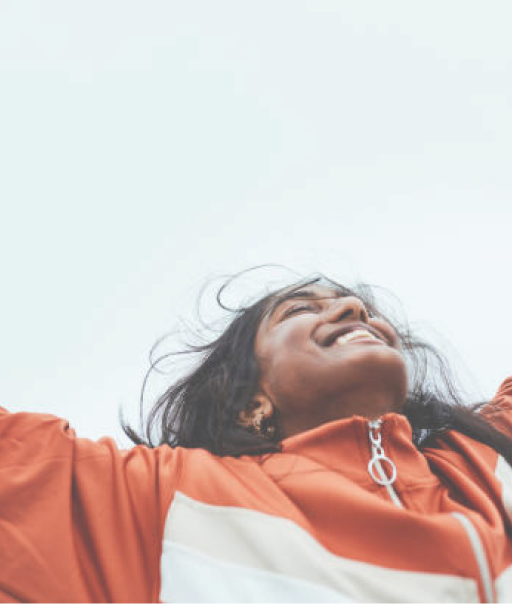 A person in an orange jacket raises their arms against a cloudy sky, exuding a sense of freedom and joy.