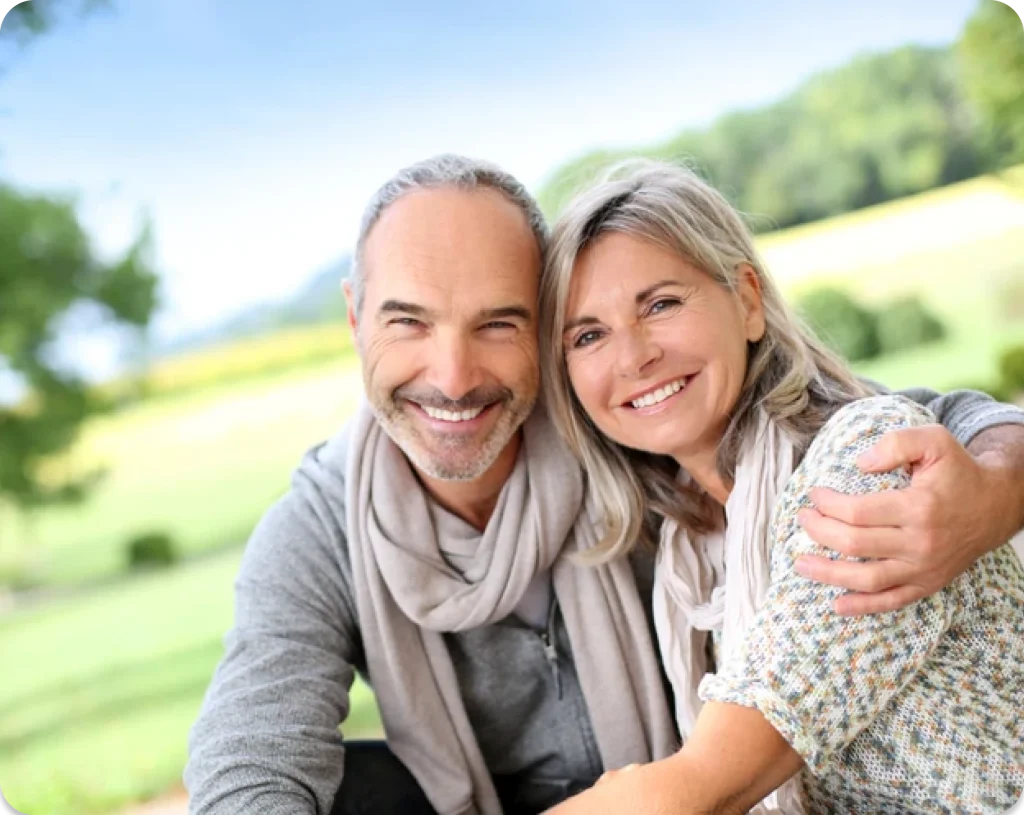 A smiling couple embraces in a sunny outdoor setting, surrounded by greenery and a blurred landscape in the background.