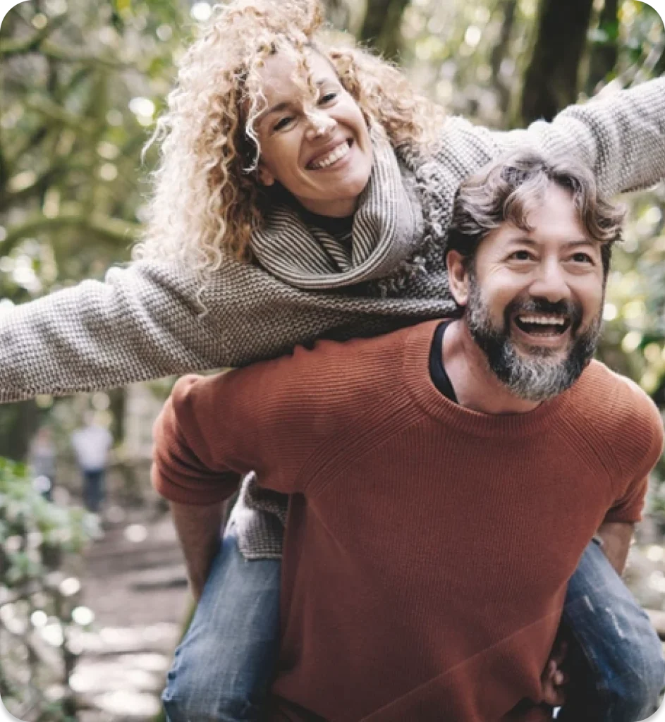 A person playfully gives a piggyback ride through a lush, green forest, surrounded by trees and vibrant foliage. A joyful person with curly hair rides on the shoulders of another, both smiling in a lush, green forest setting.